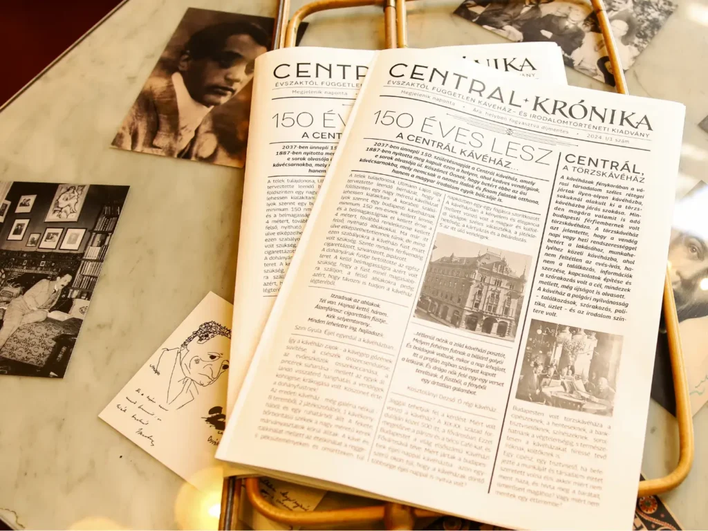 Copies of the Centrál Krónika café newspaper on a marble table with old photos of early 20th-century Hungarian writers